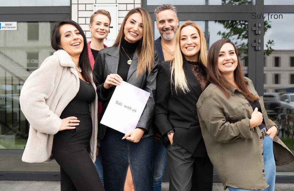 Cały zespół Savfron, wszyscy uśmiechnięci. Prezes trzyma teczkę z logo Sav with Us promując autorski program pomocowy.
